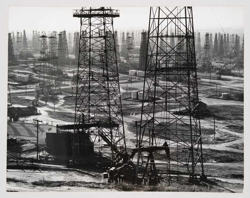 Rows of tall oil derricks and pumpjacks spread across a large, dusty oil field.