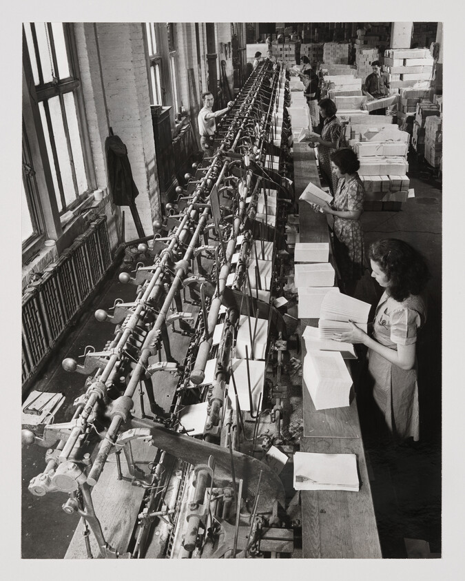 Women standing along a long assembly machine folding and stacking paper into neat piles.