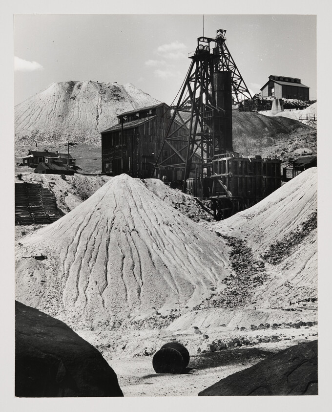A black and white photograph of an industrial mining site with a headframe and associated buildings. Large piles of excavated material dominate the foreground, with a prominent conical mound in the center. An old, rusted barrel lies on the ground to the left. The sky is clear, suggesting a dry, dusty environment.