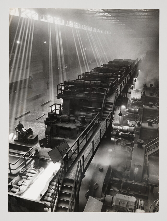 Large steel mill machinery shaping a glowing hot metal slab as sunlight streams through high windows.