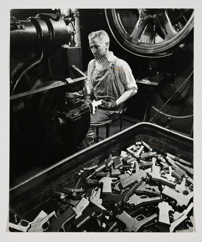 Factory worker inspects a metal part while seated beside machinery and a bin of cast components.