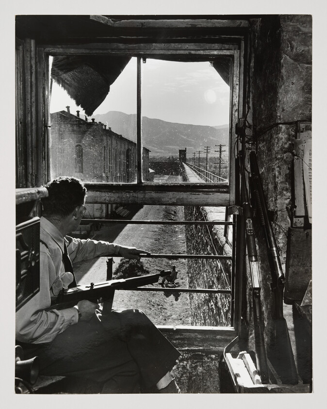 A guard with a rifle watches prisoners in a sunlit yard from a high windowed tower.