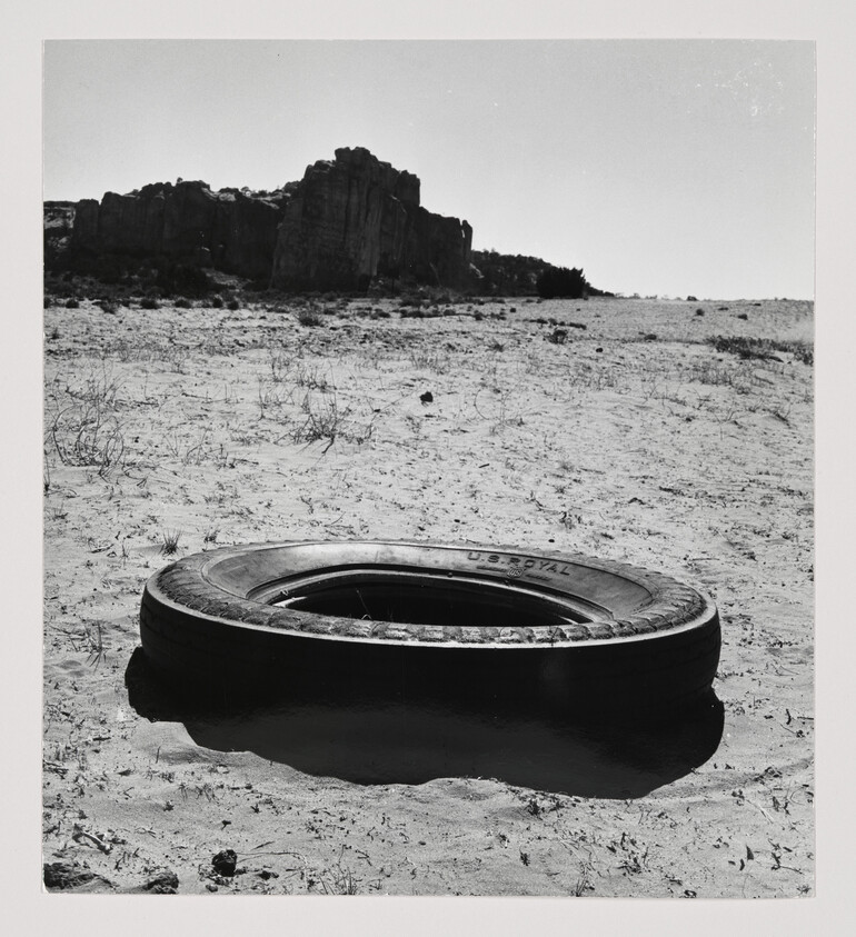 A discarded tire lies partially sunken in sandy desert with rocky cliffs in the distance.