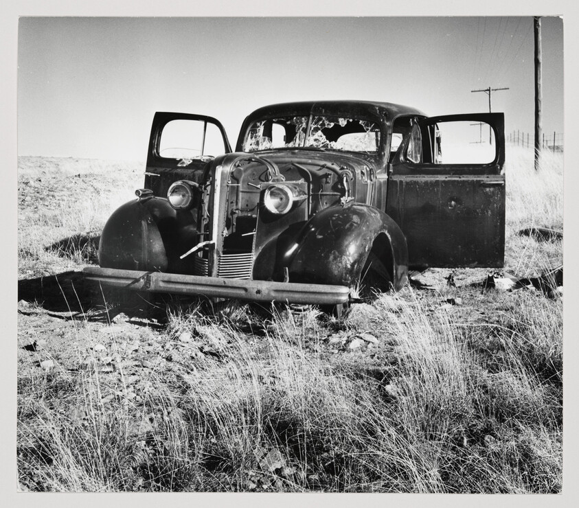 An abandoned, battered vintage car with broken windows and open doors sits in dry grass.