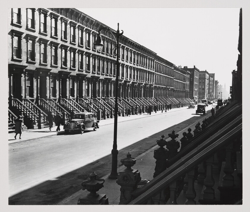 A black and white photograph capturing a sunny day on a street lined with uniform row houses featuring stoops with iron railings. A vintage car is parked on the side of the street, and a few pedestrians are visible walking on the sidewalk. The perspective is from an elevated vantage point, possibly a nearby staircase, with ornate balusters partially framing the bottom right of the image. Shadows cast by the street lamps and buildings create a contrast of light and dark across the scene.