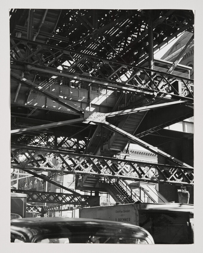Steel elevated train tracks and stairways cross above cars on a busy urban street.