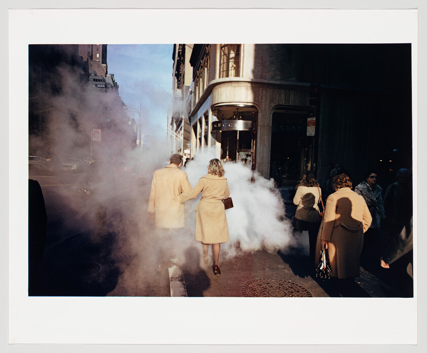 A couple in matching tan coats walk into a cloud of steam on a city sidewalk.