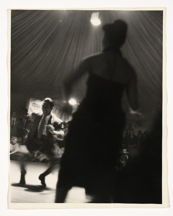 A silhouetted woman watches an energetic man dancing under a tent while a crowd looks on.