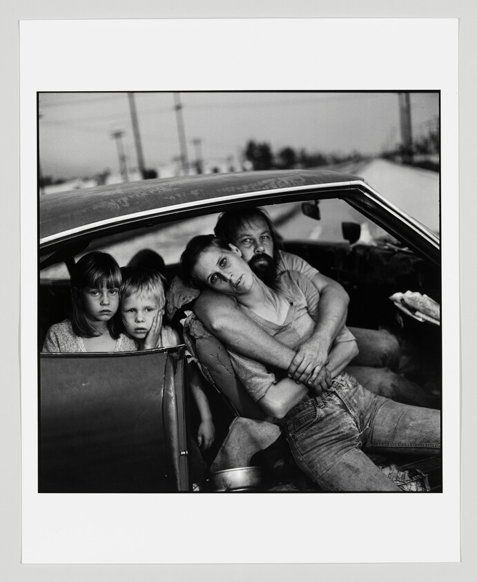 A tired-looking family huddles in a car, parents embracing while two children stare out.
