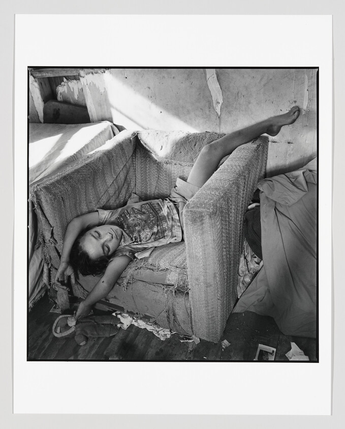 Black and white photograph of a person lying on a worn-out couch with exposed stuffing, in a relaxed pose with one arm draped over their forehead and one leg propped up against the back of the couch. Sunlight filters into the room, casting shadows on the wall and couch. The scene suggests a moment of rest in a dilapidated interior setting.