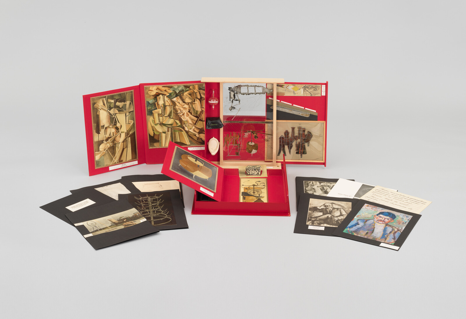 A collection of various artworks and artifacts displayed on a neutral background. The centerpiece is a red, open box containing multiple compartments, each holding different items such as a small sculpture, a bottle, and several framed pieces of art. Surrounding the box are additional framed artworks and photographs laid out, some with descriptive text beneath them. The collection appears to be part of an exhibition or a curated set.