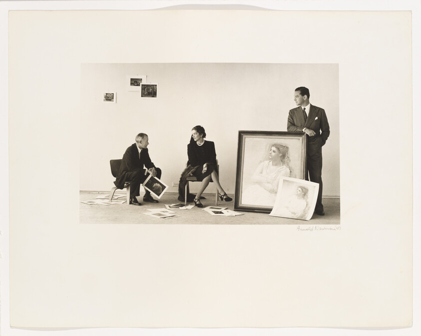 Two seated people examine photographs on the floor while a man stands holding a large framed portrait.