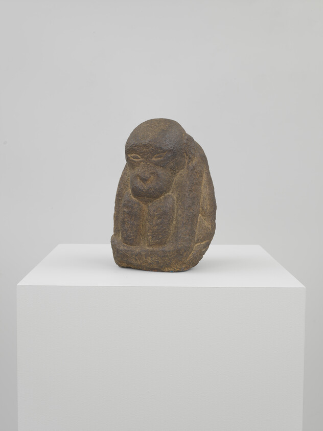 Small carved stone monkey sculpture sits on a white pedestal against a plain wall.