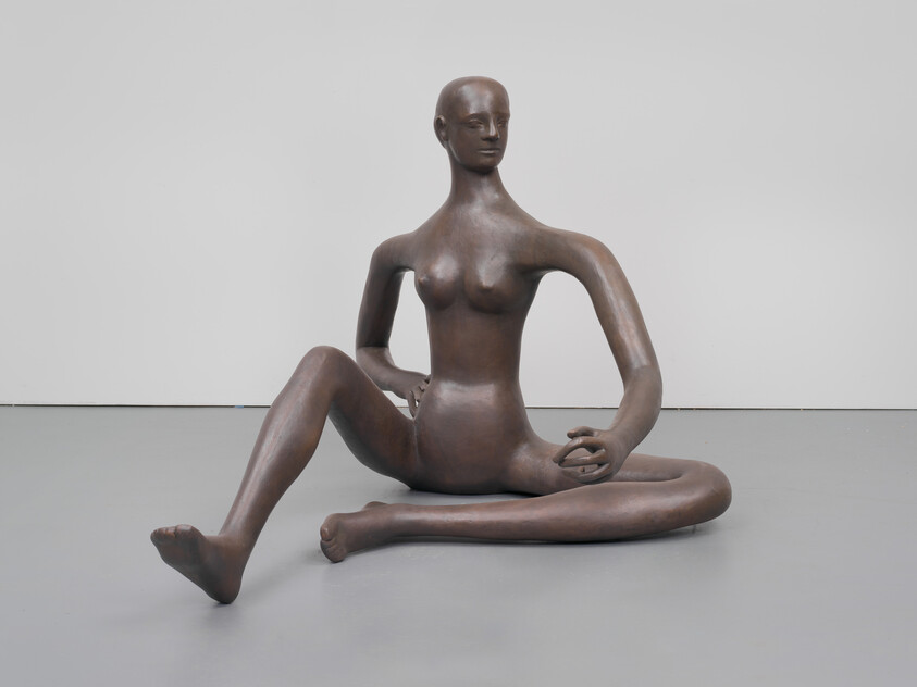 Large bronze sculpture of a seated female figure with elongated limbs and a relaxed pose.