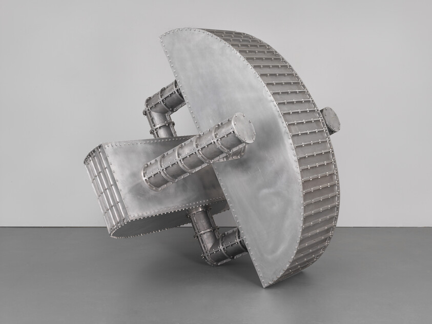 A large riveted metal sculpture with cylindrical supports leaning diagonally on a gallery floor.