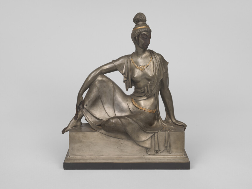 Bronze statue of a seated woman in draped clothing with gold-accented jewelry on a rectangular base.
