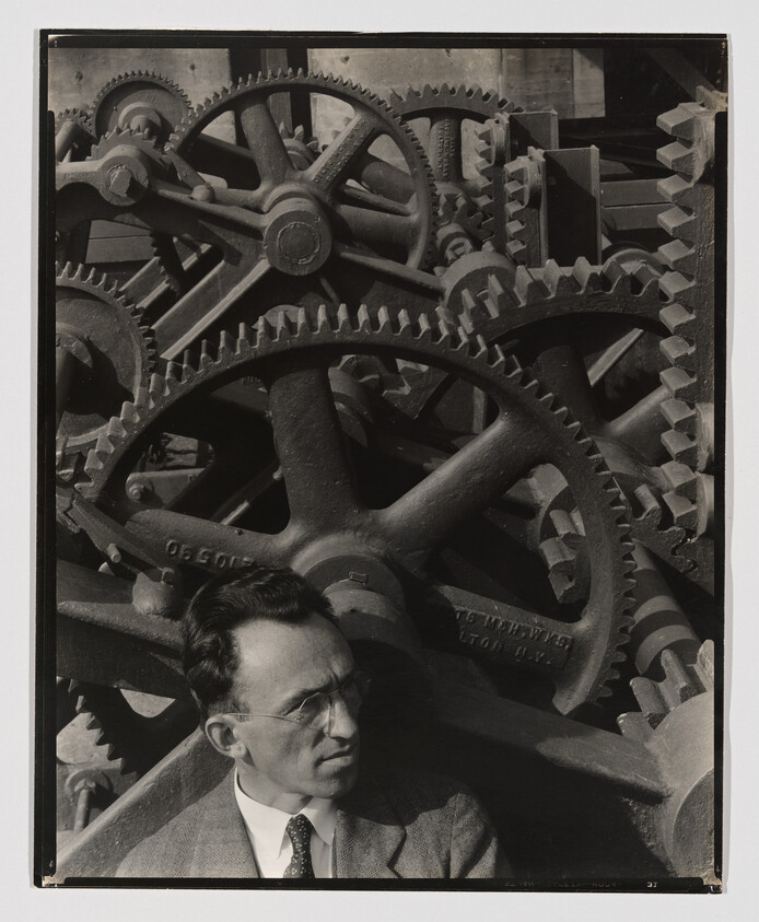 Black and white photograph of a man in a suit and glasses sitting in front of a large assembly of interlocking industrial gears and cogs. The machinery dominates the background, creating a complex pattern of mechanical parts, while the man appears contemplative.