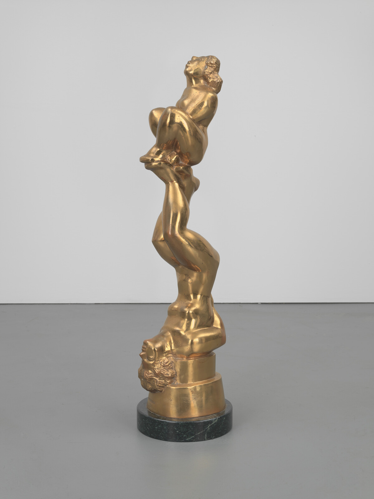 A tall gold sculpture of three stacked human figures twisting on a round dark base.