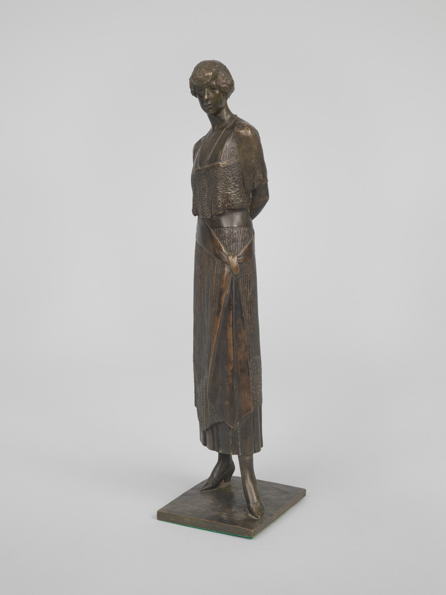 Bronze sculpture of a slender woman standing with hands behind her back wearing a long dress.
