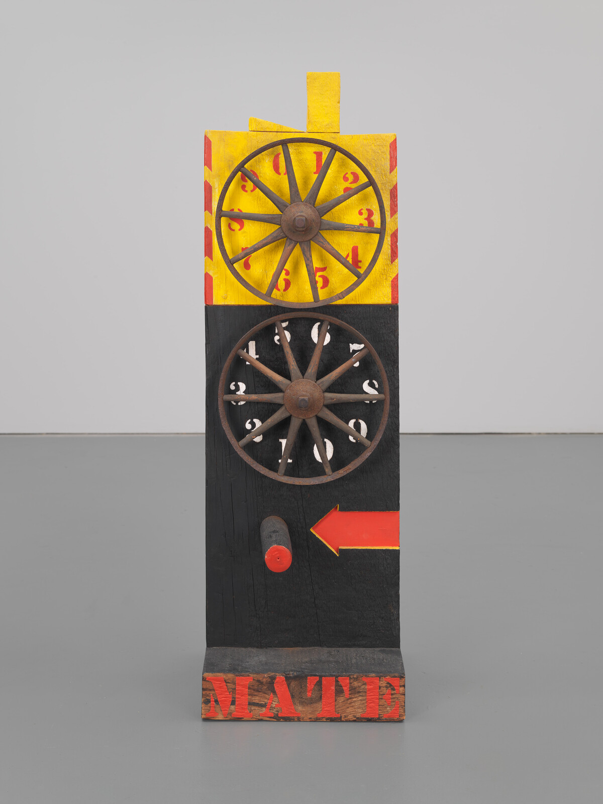Wooden sculpture with two wheels, numbers, and an arrow. The word "MATE" is painted at the bottom.