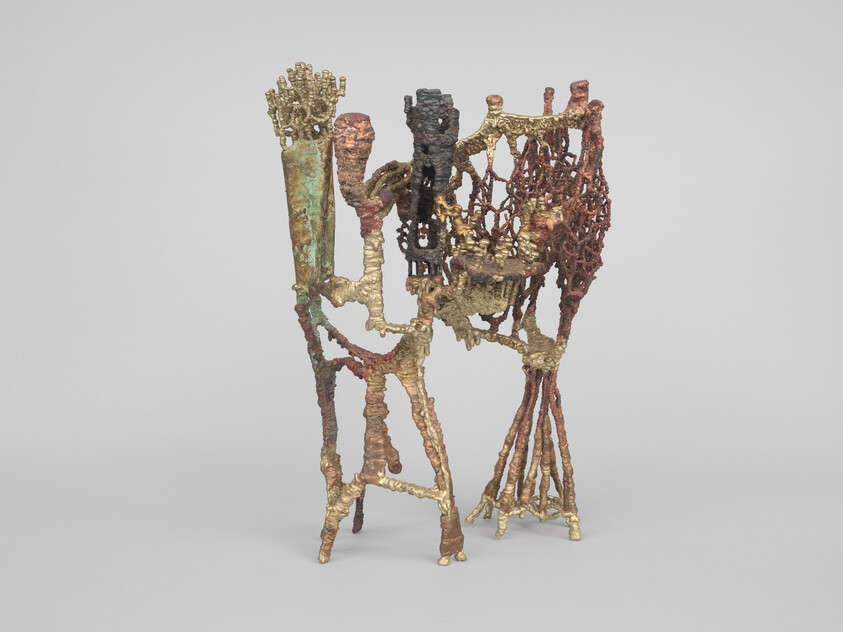 Abstract metal sculpture with intertwined lattice-like forms and uneven legs standing on a gray surface.