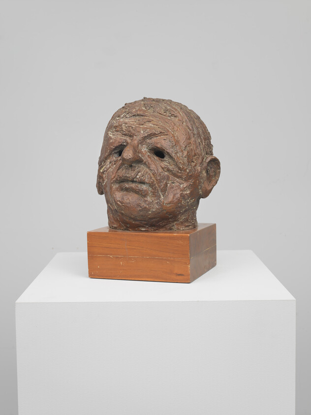A rough-textured sculpted male head bust sits on a wooden base atop a white pedestal.
