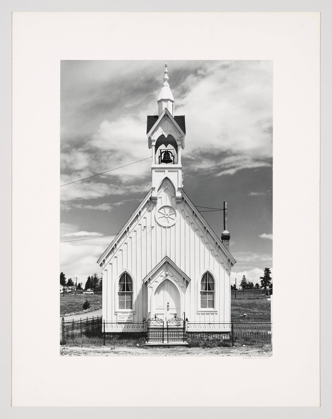 Black and white photograph of a traditional wooden church with a steeple and bell tower, set against a cloudy sky. The church features Gothic-style windows and a prominent front entrance with steps leading up to double doors. A simple metal fence surrounds the property, and the background shows a rural landscape with sparse trees.