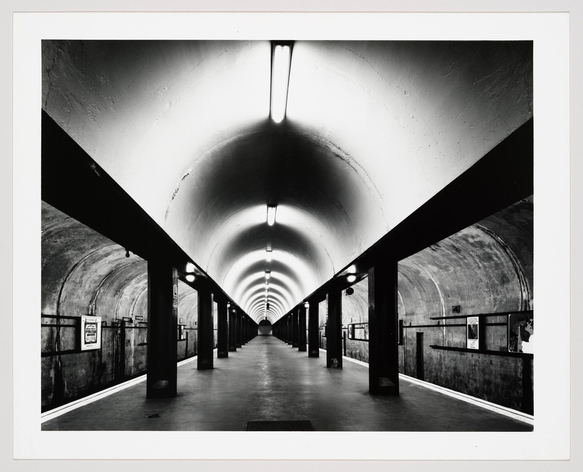 A black and white photograph of a long, empty subway station with a curved ceiling and a series of columns on either side. Fluorescent lights line the ceiling, casting a bright glow and creating a vanishing point in the distance.