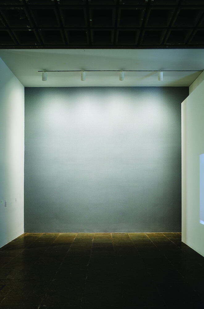 Empty gallery wall illuminated by four ceiling spotlights above a dark tiled floor.