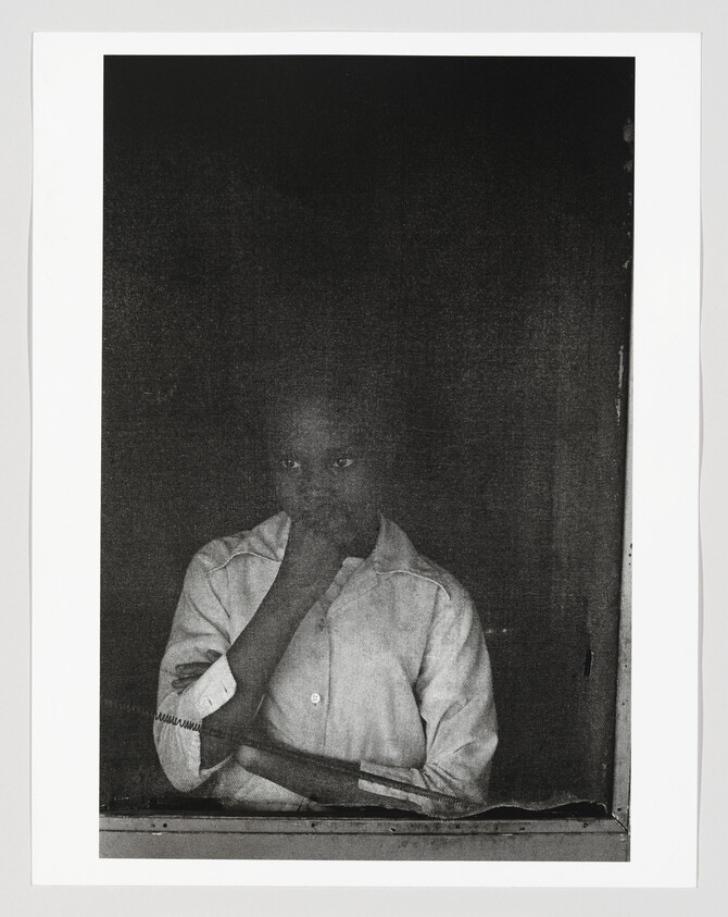 A young boy stands at a window with his hand to his mouth, looking thoughtful.