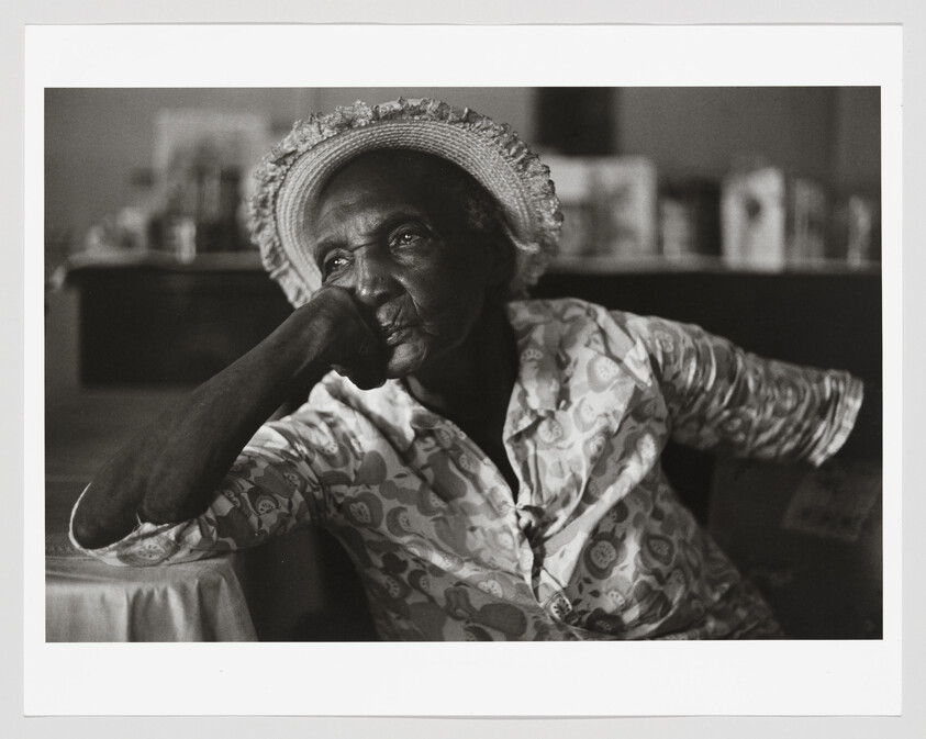 Elderly woman in patterned shirt and straw hat rests her chin on her hand, looking thoughtful.