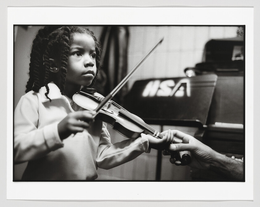 A young girl concentrates while playing a violin as an instructor guides her hand.