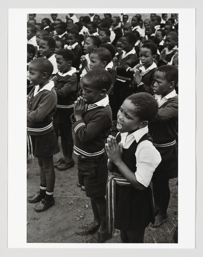 Young schoolchildren stand in rows with hands clasped together, appearing to pray or sing.