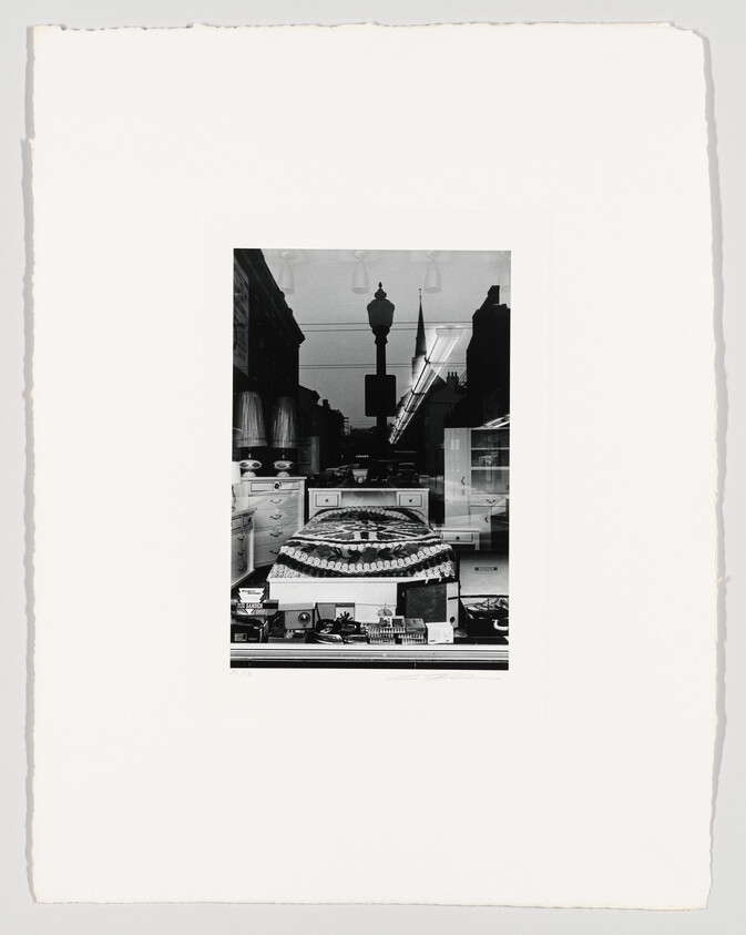 Black and white photograph depicting a street scene reflected in a shop window, showcasing a variety of small items on display, with buildings and street lamps visible in the reflection. The image is centered on a textured white background.