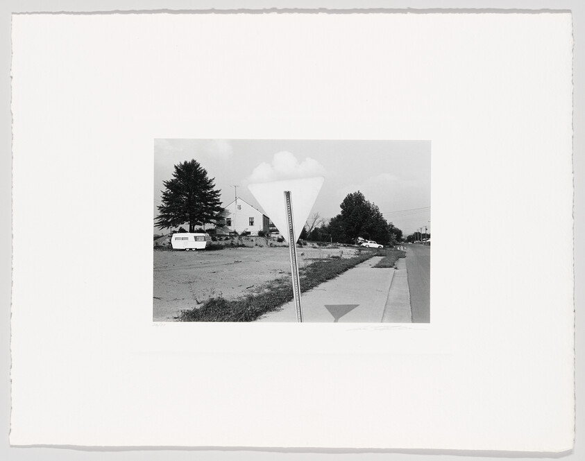 A faded triangular road sign stands beside a sidewalk in front of a house and parked trailer.