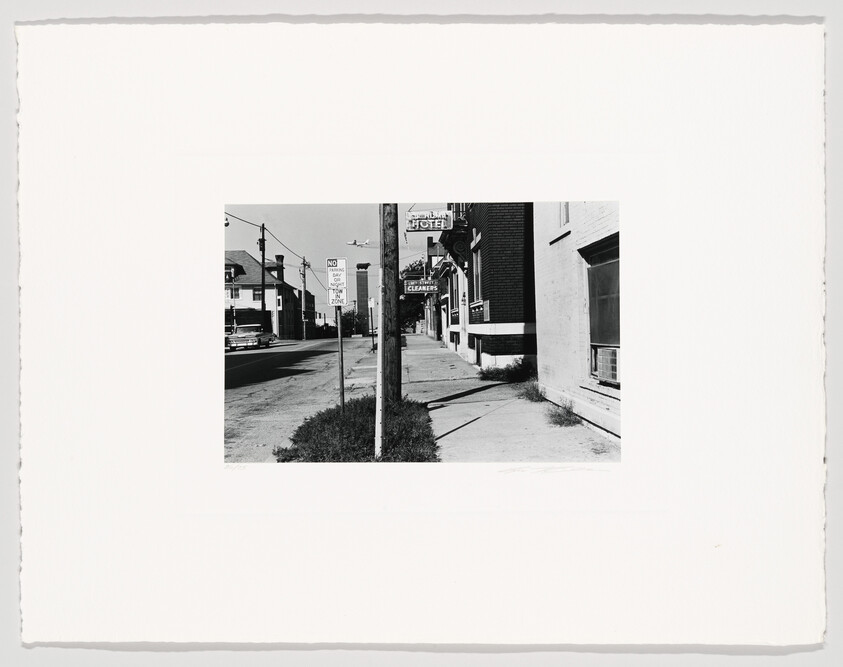 Black and white photograph depicting a mid-20th-century street scene with vintage cars parked along the curb, various storefronts including a hotel and a cleaners, and a "NO PARKING" sign prominently displayed in the foreground. The image has a white border typical of old photographs.