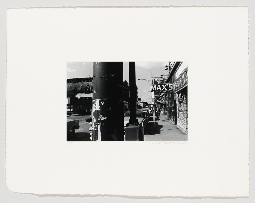 A city sidewalk with a large pole in the foreground and a storefront sign reading MAX'S.