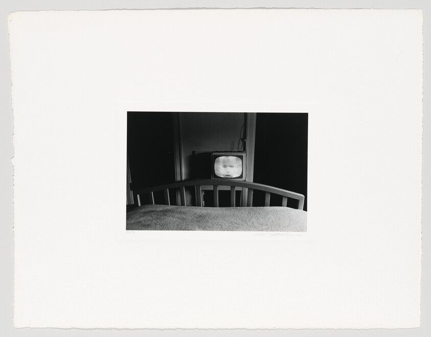 A black and white photograph depicting a vintage television set at the foot of a bed, displaying a blurry image of a face on its screen. The photo is centered on a white background with rough edges, suggesting it is a physical print.