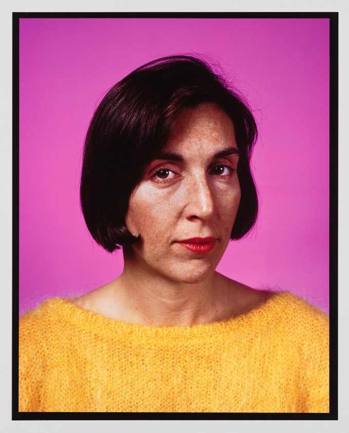 A portrait of a woman with short dark hair, wearing a bright yellow sweater, against a vivid pink background. She has a neutral expression on her face and is looking directly at the camera.