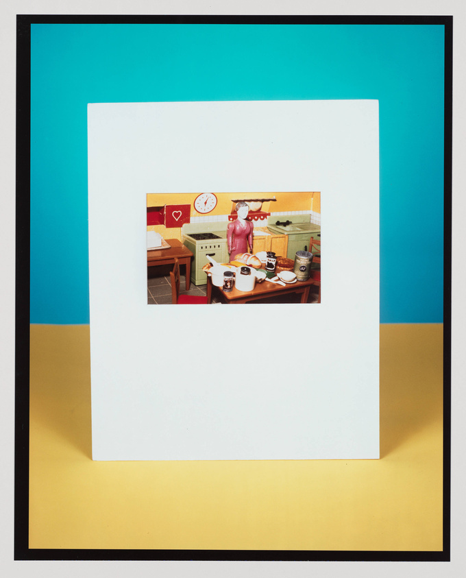 A photograph within a photograph, featuring a vintage kitchen scene with a pink mannequin figure standing at the center. The inner image is bordered by a white frame, which is set against a bright turquoise background at the top and a yellow one at the bottom, with a black border surrounding the entire composition.