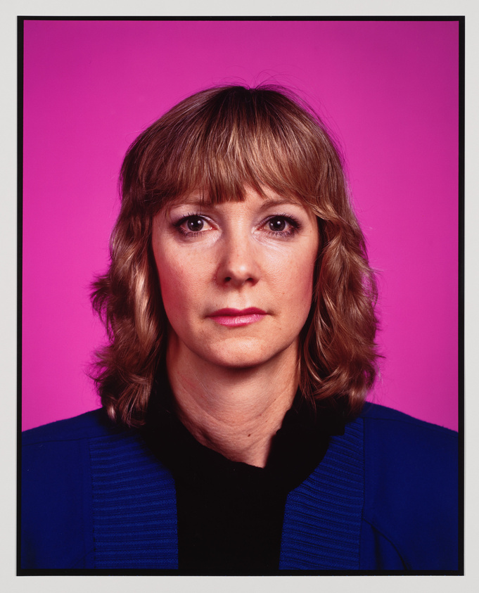 A portrait of a woman with shoulder-length wavy hair, looking directly at the camera with a neutral expression. She wears a black top with a blue jacket, and the background is a vibrant pink.