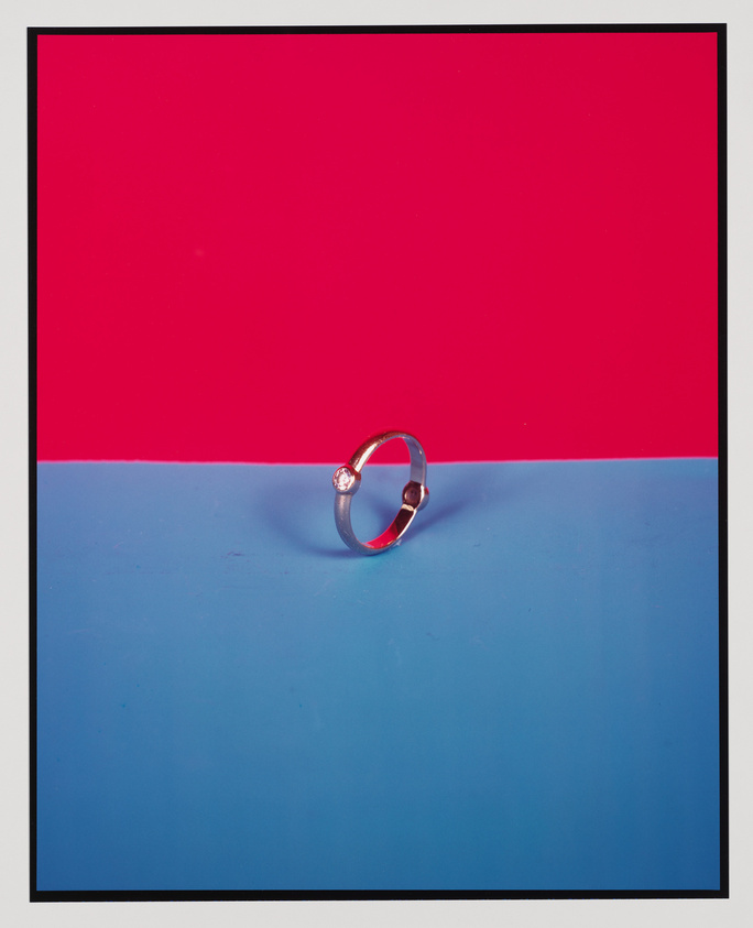 A silver ring with a single gemstone is centered on a surface that is split between a bright red upper half and a blue lower half, creating a striking contrast. The image is framed with a white border.