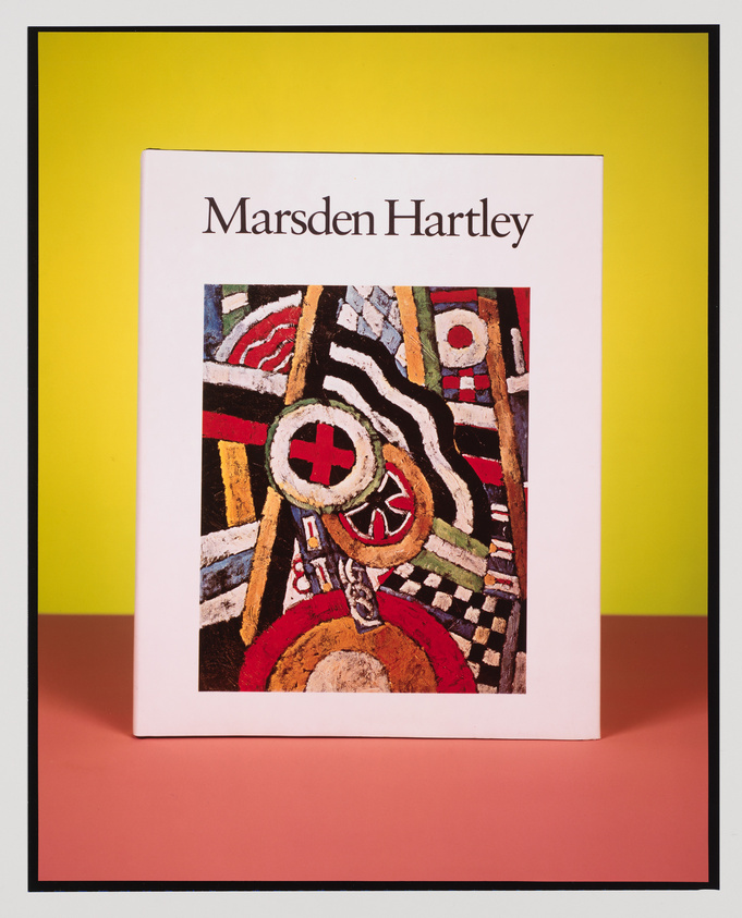 A book about Marsden Hartley is displayed upright against a yellow background with a pinkish surface beneath. The book cover features a colorful abstract painting with bold patterns and shapes, including circles and checkered designs.