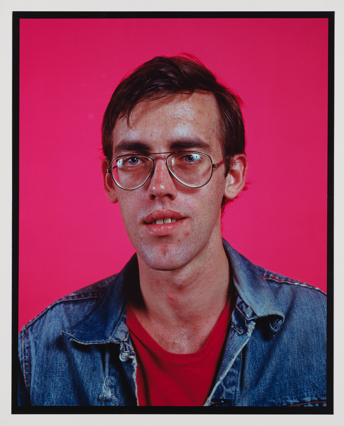 A portrait of a young man with short brown hair and glasses, wearing a denim jacket over a red shirt. The background is a vibrant pink, and the image is framed with a black border.