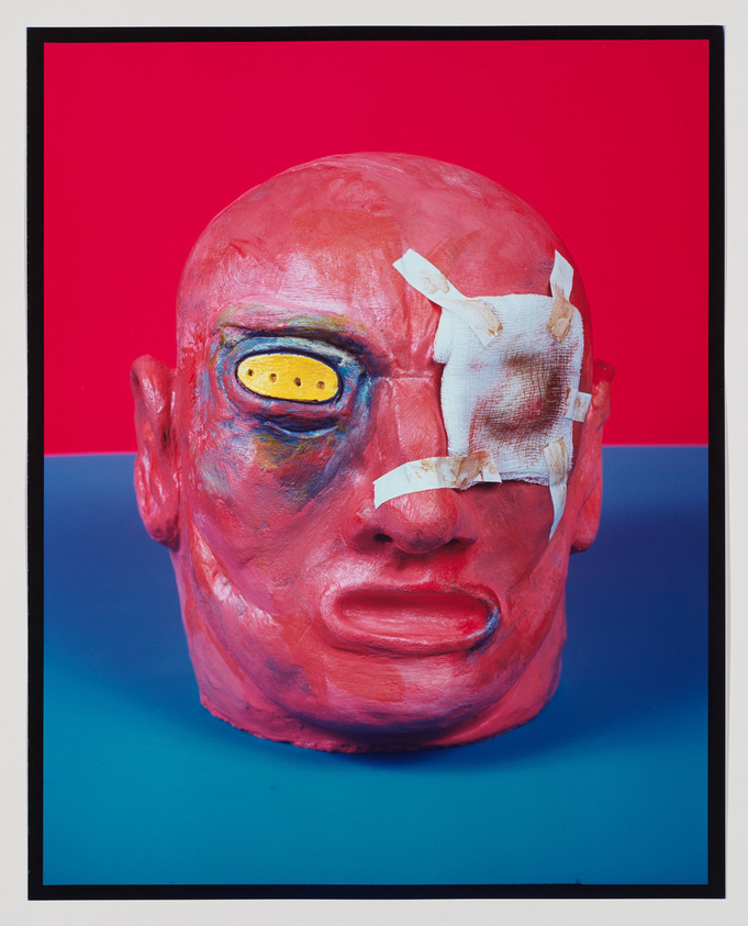 A vividly colored artwork featuring a sculpted head with exaggerated features against a red and blue background. The head is painted red with one eye covered by a bandage and the other eye depicted as a yellow oval with black dots. The sculpture has a textured surface and is framed within a black border.