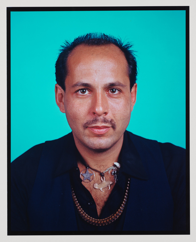 A portrait of a man with a mustache and slicked-back hair, wearing a dark shirt and a necklace with large beads and various pendants, against a bright turquoise background.