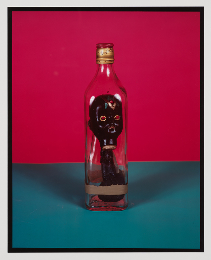 A clear glass bottle with a golden cap stands against a pink and blue background. Inside the bottle, there is a dark figurine with facial features, resembling a doll's head, and a bow on its head. The figurine appears to be submerged in a dark liquid at the bottom of the bottle.