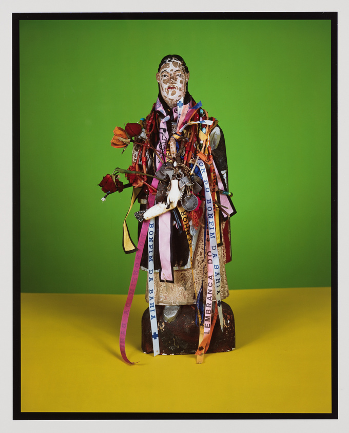 A colorful and eclectic sculpture composed of various objects and materials, including ribbons, flowers, and a mask, against a green background with a yellow floor.