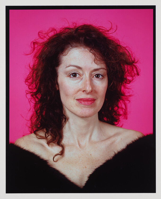 Woman with curly dark hair and bare shoulders posing against a bright pink background.