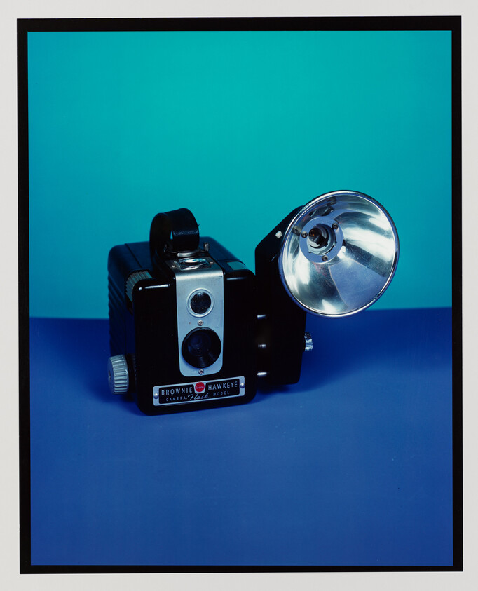 A vintage Brownie Hawkeye box camera with a large attached flash reflector on a blue background.